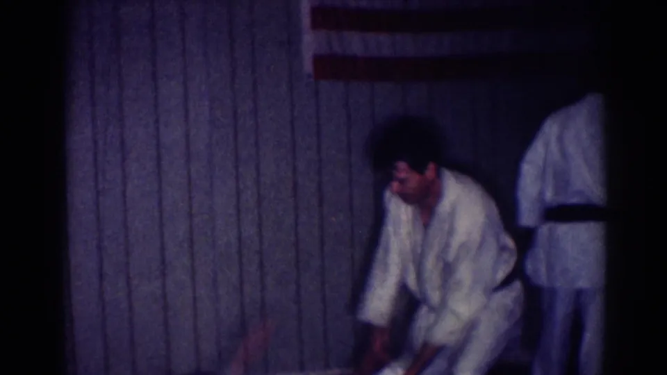 A karate practitioner bends forward near the floor as another person in a white gi is partly visible at the edge of the frame.