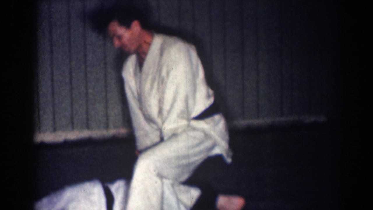 Man in a white gi and dark belt leans over another person on a mat in a dim dojo.