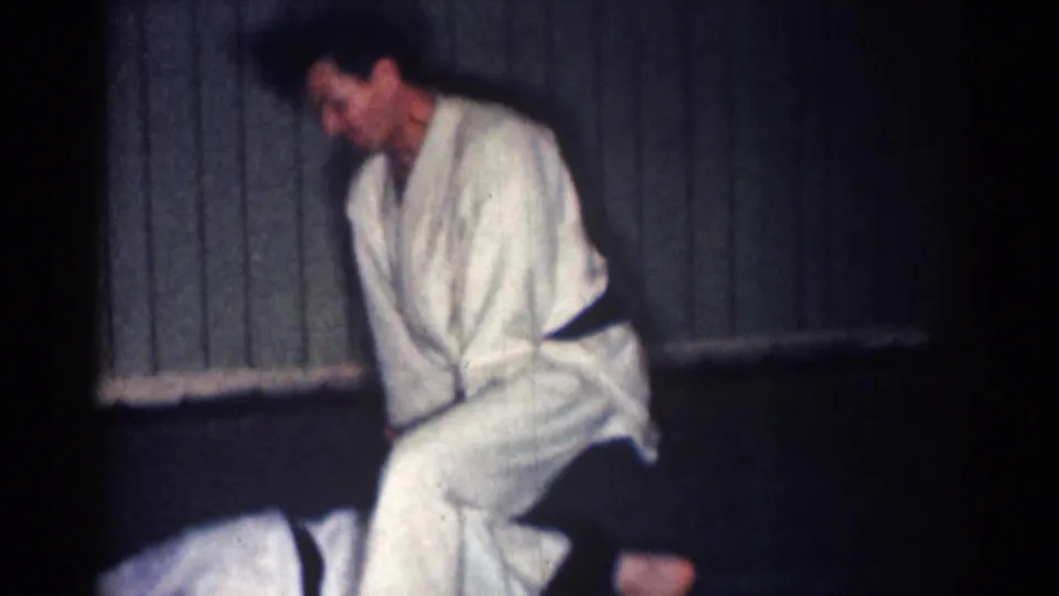 1967 Los Angeles 8mm frame showing a man in a white gi and dark belt bending over an opponent on a mat inside a paneled dojo.