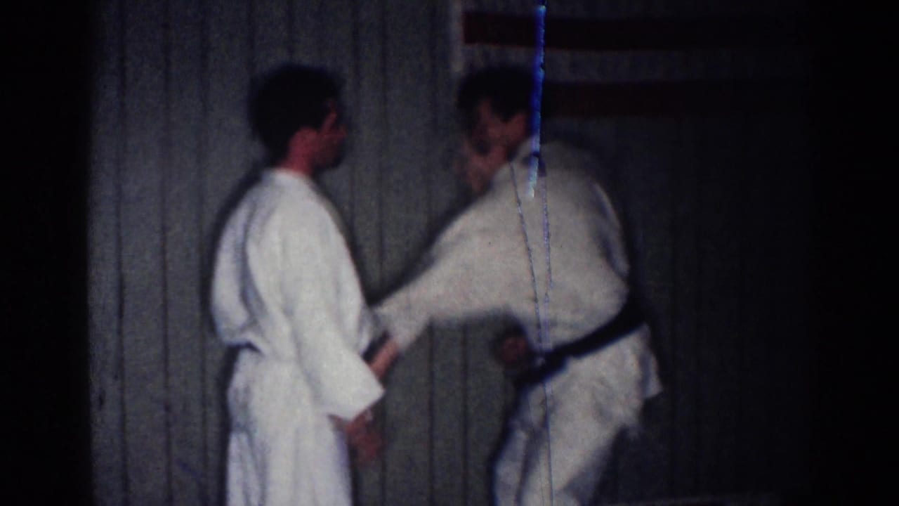 Two men in white gis face each other during a karate exchange in front of paneled walls.