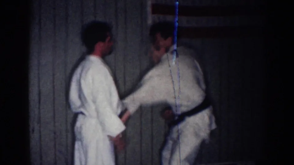 1967 Los Angeles 8mm frame of two men in white gis and dark belts facing off during a karate match in a dim paneled room.