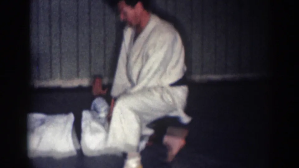 1967 Los Angeles 8mm frame showing a karate participant standing over an opponent on the mat inside a small paneled dojo.