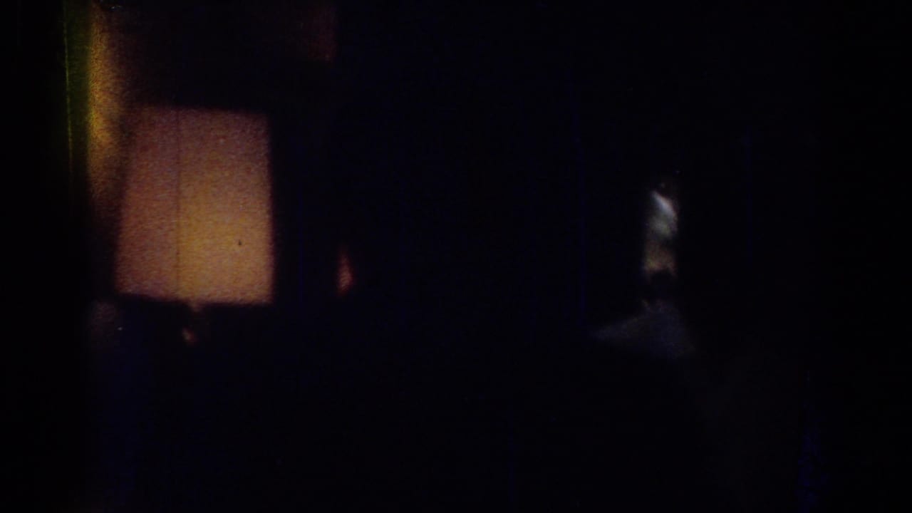 Dark interior with a warm lamp glow and indistinct shadowed forms.