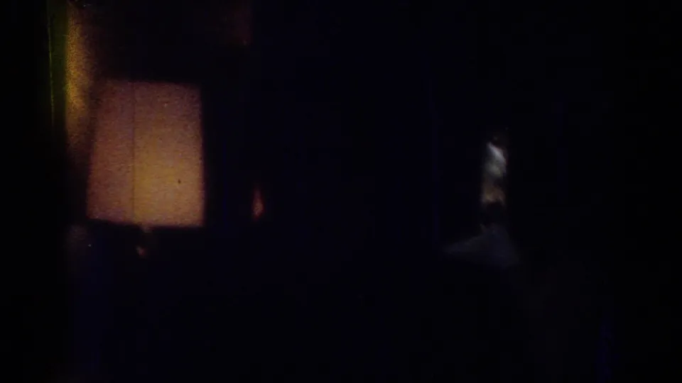 Low-light 1967 Los Angeles home-movie frame with a warm lamp at left and indistinct figures or furnishings in shadow.