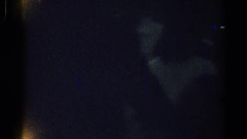 Underexposed 8mm frame from the Los Angeles party clip, showing faint reflections and possible human silhouettes in a dark interior.