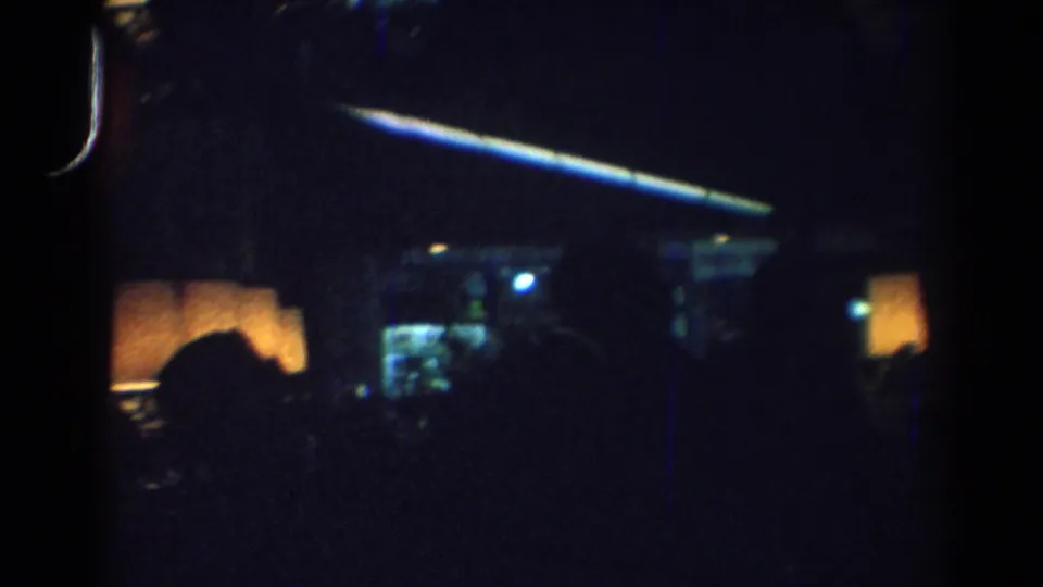 1967 Los Angeles 8mm frame showing a dim bar interior with seated silhouettes, amber lamps, and a blue ceiling strip.