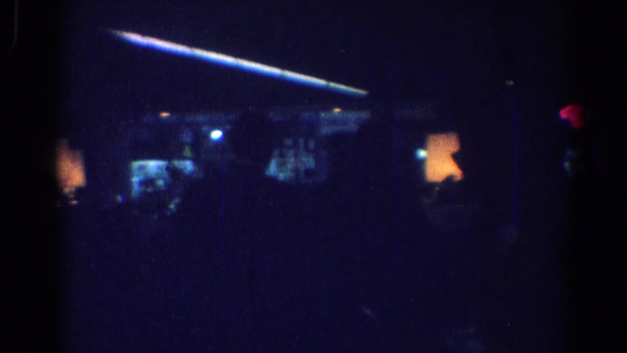 Dim interior with blue ceiling light, warm lamps, and shadowed people seated below.