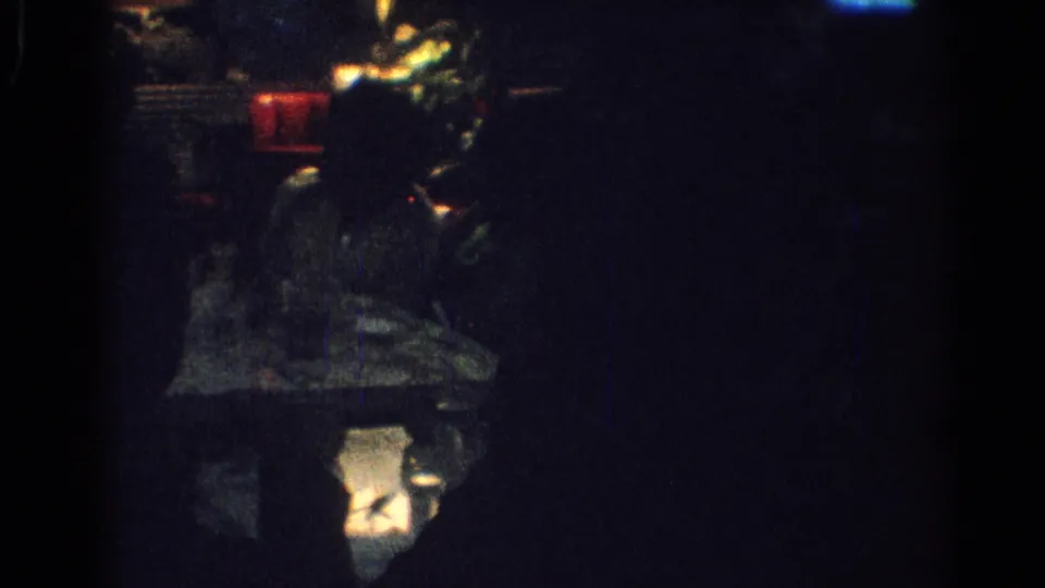 1967 Los Angeles 8mm frame showing a shadowed seated woman in a low-lit dining or lounge interior with faint red highlights.