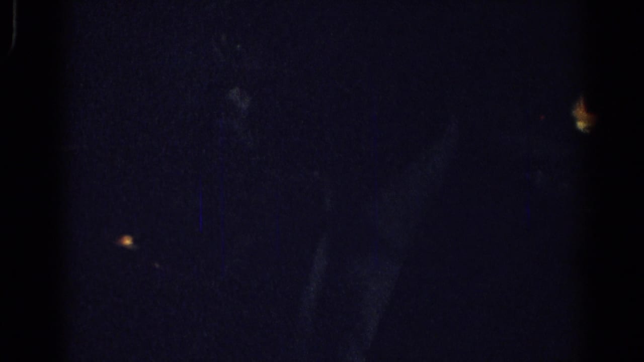 Very dark frame with a faint figure and small points of light in the background.