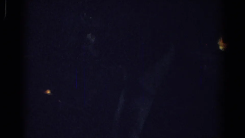 Low-exposure 1967 Los Angeles home-movie frame with a faint seated figure and scattered interior lights in heavy shadow.