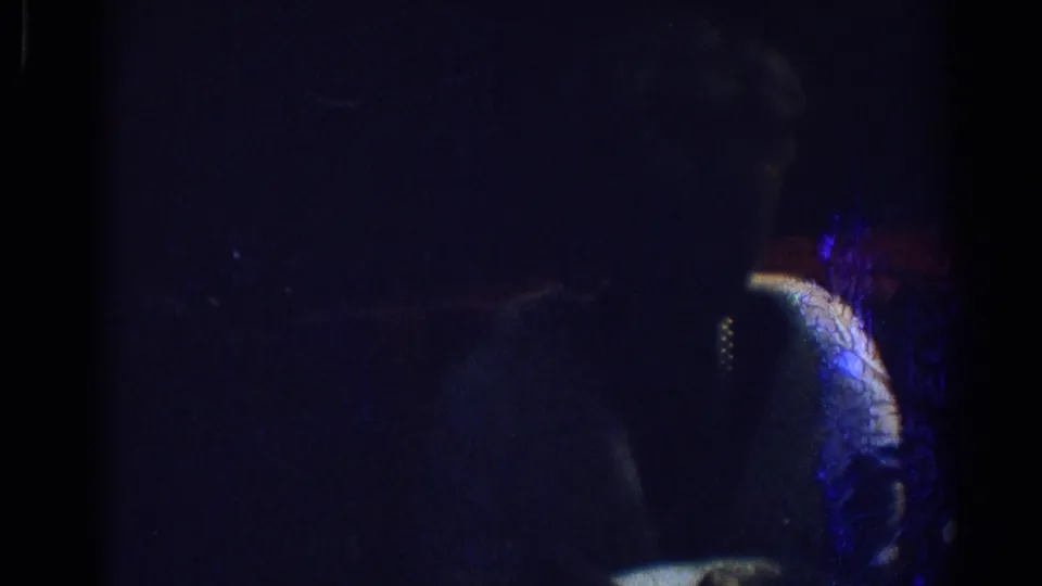 1967 Los Angeles 8mm frame of a seated woman in a pale dress or top, partially illuminated by blue and red low interior light.