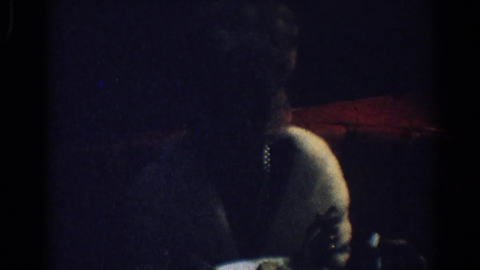 1967 Los Angeles home-movie frame showing a shadowed seated woman beside a red surface, with her shoulder and face partly lit.