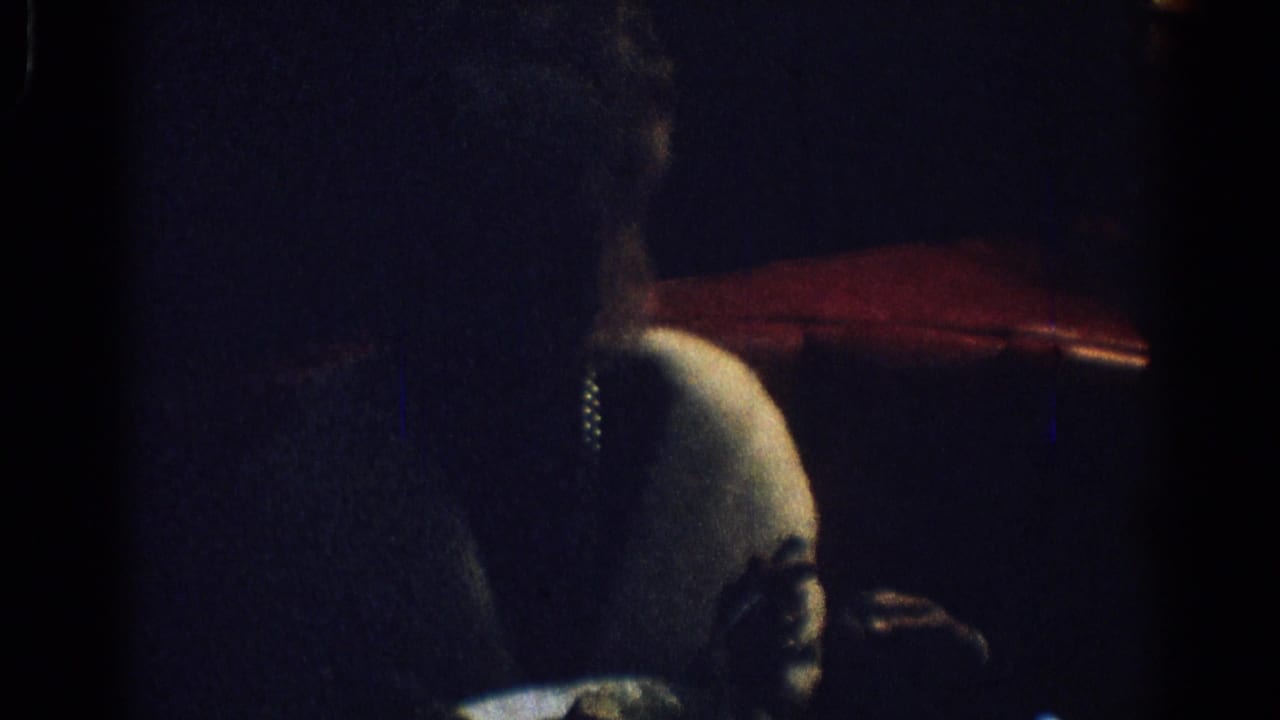 1967 Los Angeles home-movie frame showing a woman in a white fur stole seated in profile in a dark restaurant interior.
