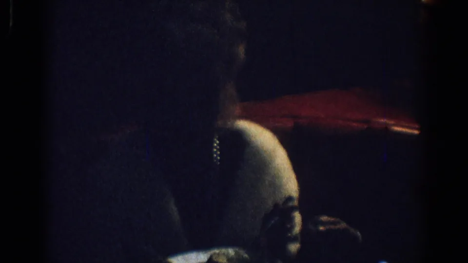 1967 Los Angeles home-movie frame showing a woman in a white fur stole seated in profile in a dark restaurant interior.