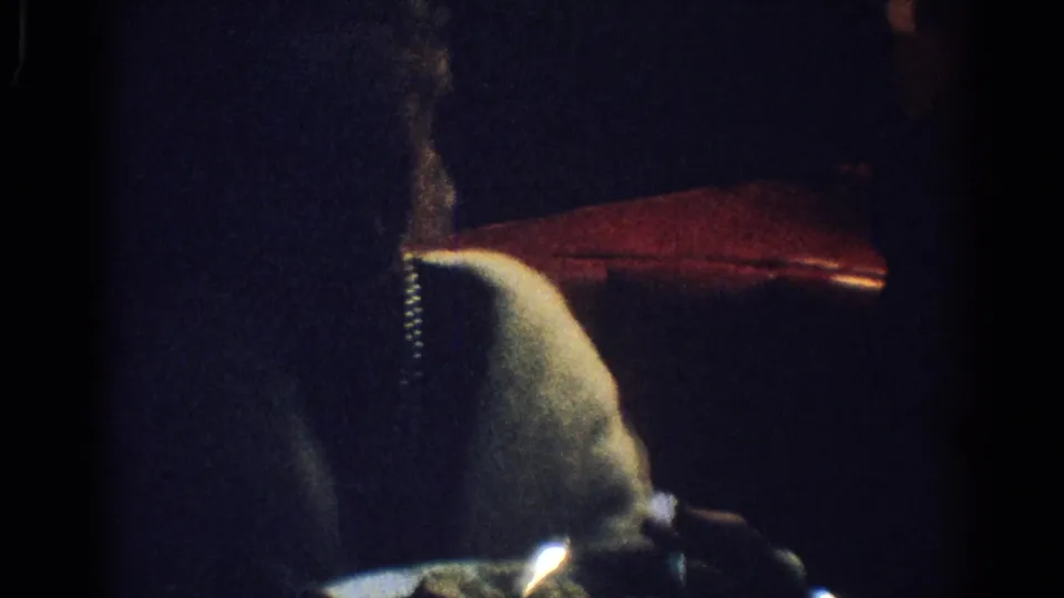 A woman in a light-colored top sits at a dim dinner table in 1967 Los Angeles, partly obscured by darkness and film grain.