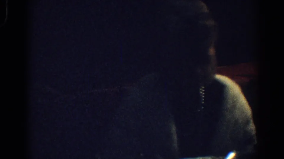 Dark 8mm frame from a 1967 Los Angeles dinner scene with a shadowed adult figure and faint table details.