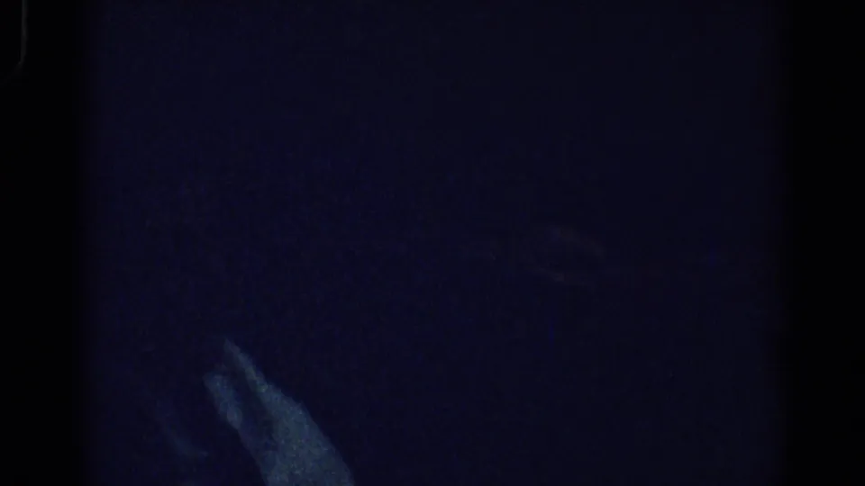 1967 Los Angeles 8mm frame showing a seated adult partly hidden by darkness, with a pale hand visible against a blue-tinted interior.