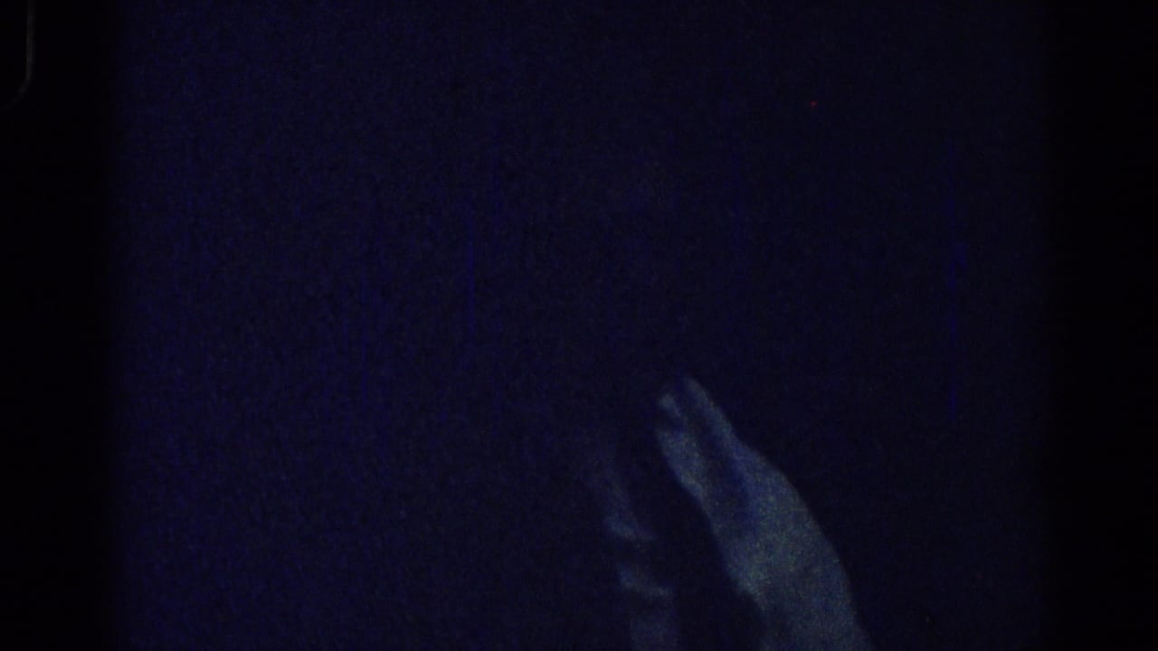A pale hand and faint seated figure appear in a dark, blue-toned interior.