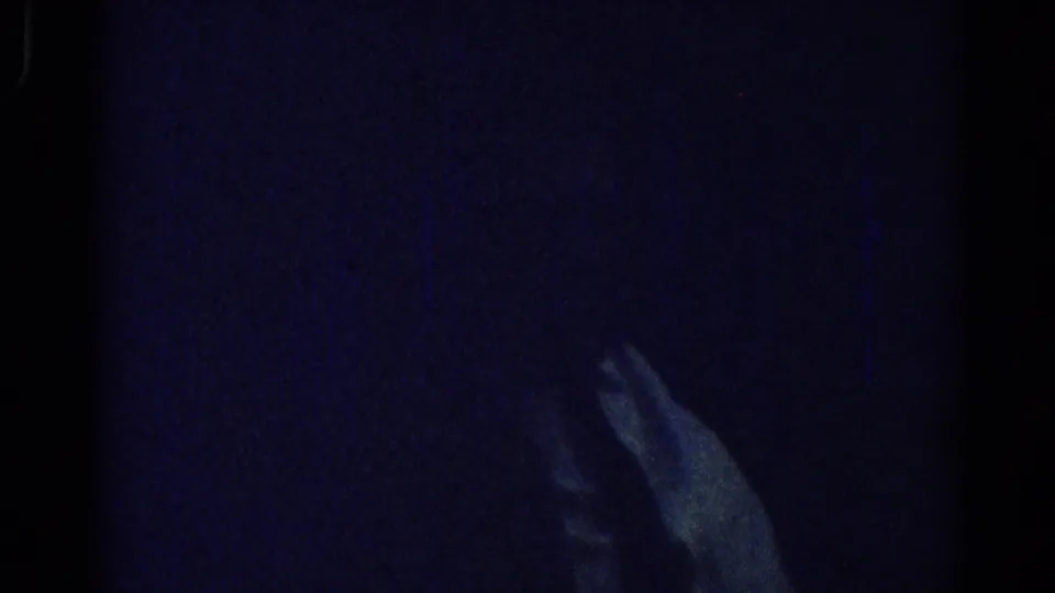 Dim 1967 Los Angeles home-movie frame with a seated adult mostly obscured and one hand visible in heavy shadow.