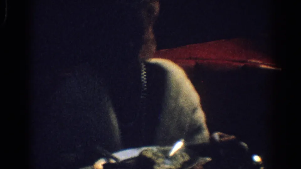 1967 Los Angeles 8mm frame showing a man in a dark suit beside a woman in a light top or stole, partly lit near a red shade-like object.