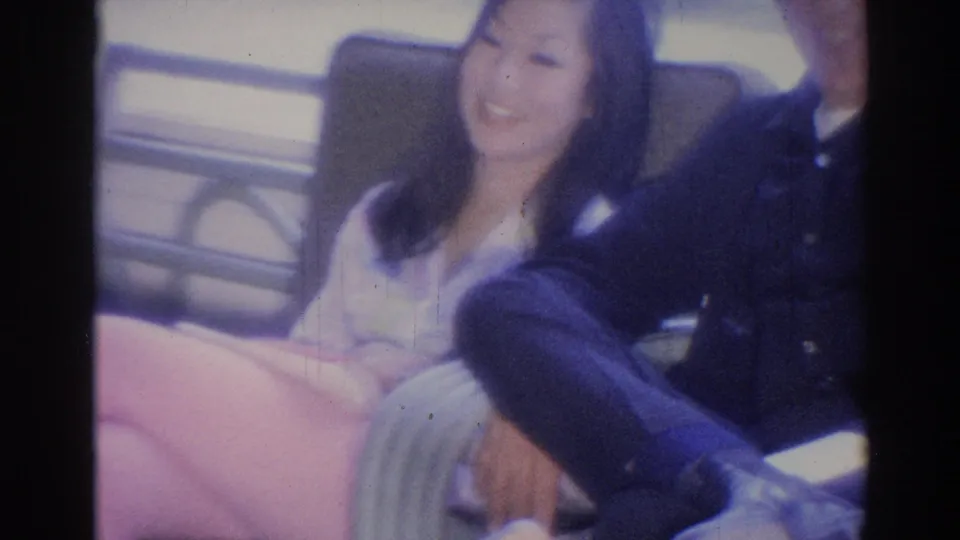 1967 Los Angeles home-movie frame of a smiling woman in light clothing seated close to a man in a dark suit on a lobby bench.