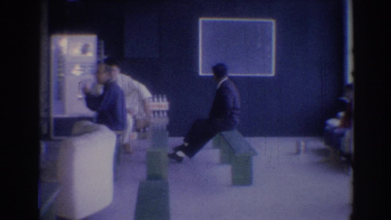 Men sit in a lobby with benches, wall art, and bottles or containers nearby.