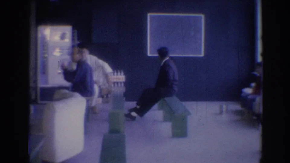 Wide 1967 Los Angeles lobby view with men seated on benches, a blue wall, framed wall art, and possible product containers in the room.