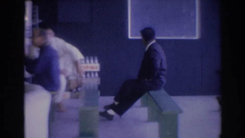 1967 Los Angeles lobby frame showing a man in a dark suit seated on a green bench as another adult moves near containers at the side.