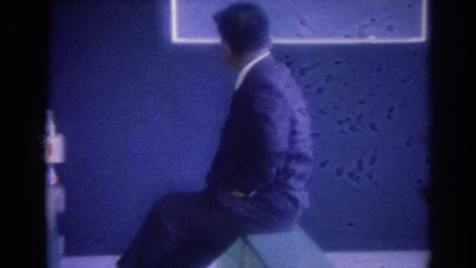 Soft 8mm frame of a suited man seated sideways on a green bench in a Los Angeles lobby, facing a blue wall and framed wall art.