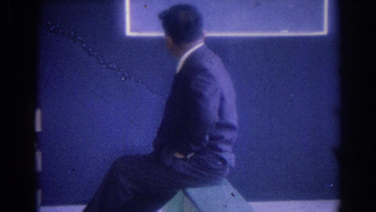 In 1967 Los Angeles footage, a man in a blue jacket is seen from behind inside a dim storefront-like interior.