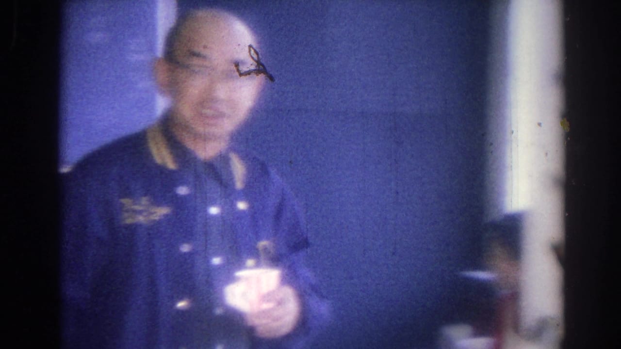 Bald man in blue jacket holding a mug near a dark wall.