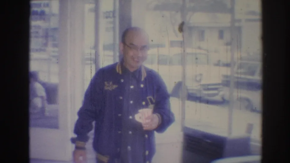 A bald man in a blue jacket holds a mug by storefront glass, with a Los Angeles street and parked cars visible outside.