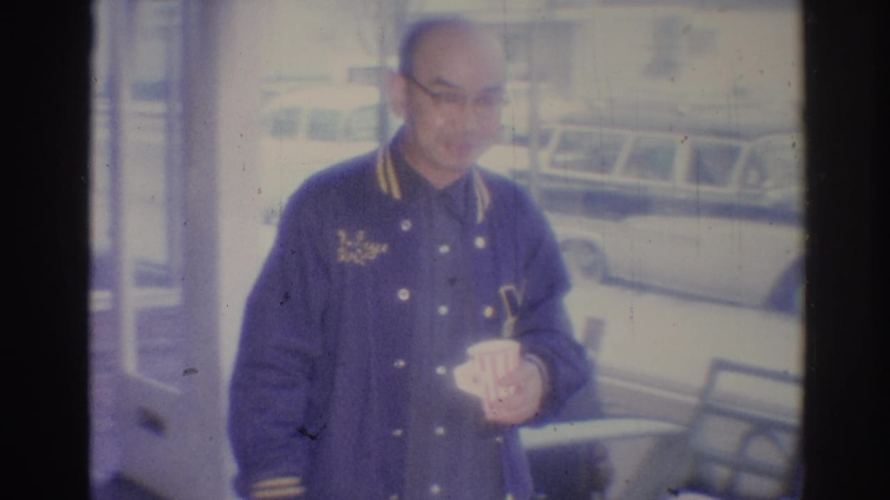 In 1967 Los Angeles home-movie footage, a man in a dark jacket stands near indoor seating while holding a striped cup, with a vehicle faintly visible beyond the bright background.