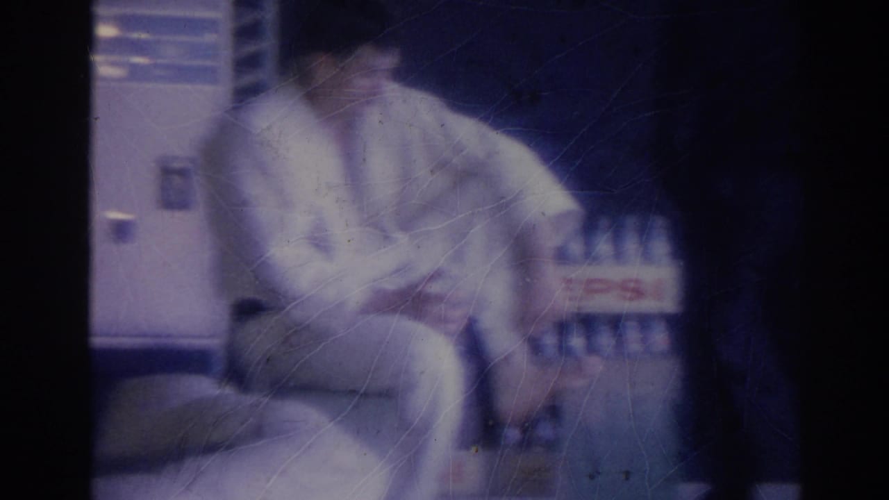 A man wearing a white gi sits in a dim interior beside a visible Pepsi bottle crate, with his face partly obscured by motion blur and soft film exposure.