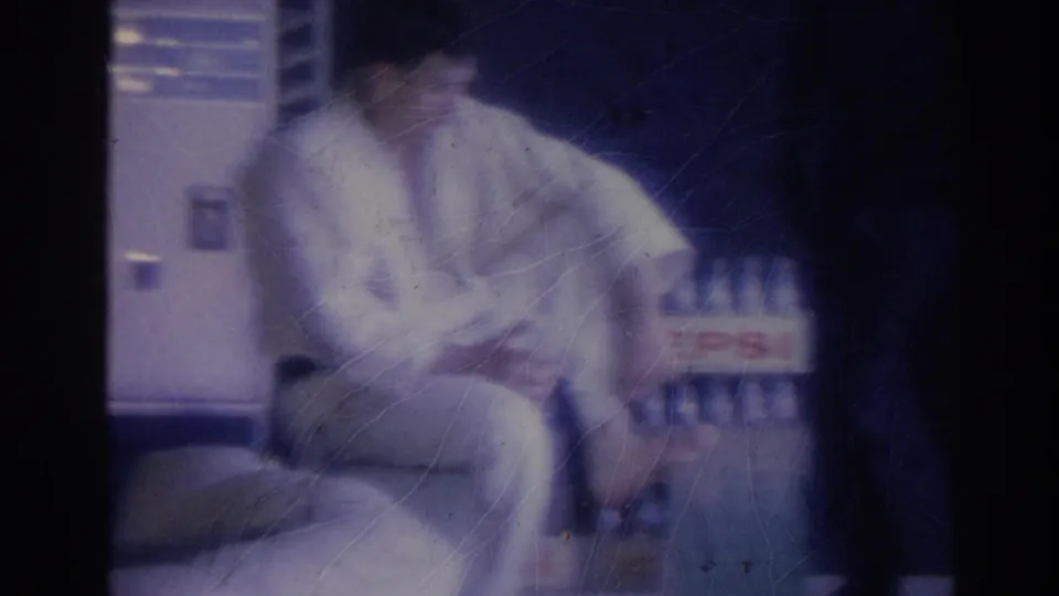 A man wearing a white gi sits in a dim interior beside a visible Pepsi bottle crate, with his face partly obscured by motion blur and soft film exposure.