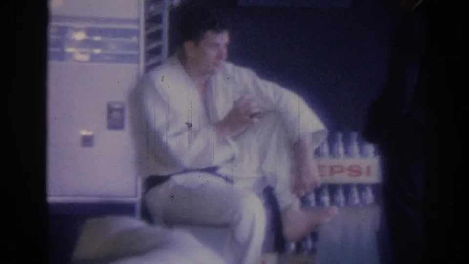 The seated man in a white gi and dark belt appears to rest with one hand near his body beside a Pepsi crate in a dim interior setting.