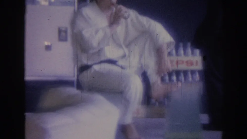A seated man in a white gi and dark belt remains beside the Pepsi crate, with the interior doorway and dark surrounding wall visible in the soft 8mm frame.