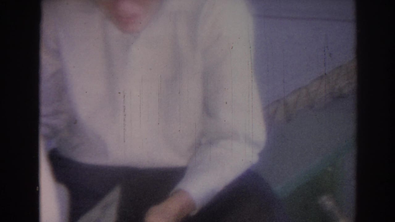 Man in a white shirt leans forward beside a wall and chair indoors.