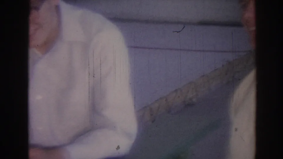 A 1967 Los Angeles home-movie frame shows a seated man in a white shirt with another person partly visible at the edge.