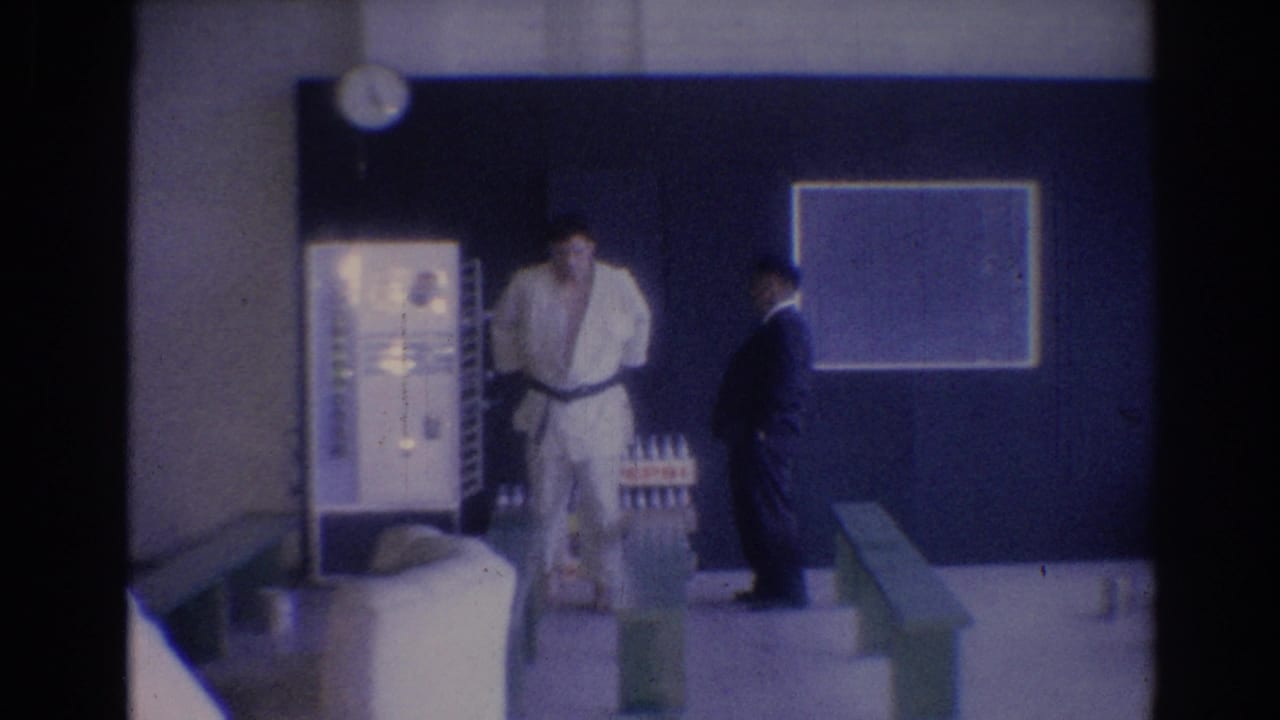 1967 Los Angeles gym frame with a judo-uniformed man near a vending machine, chalkboard, and suited man.