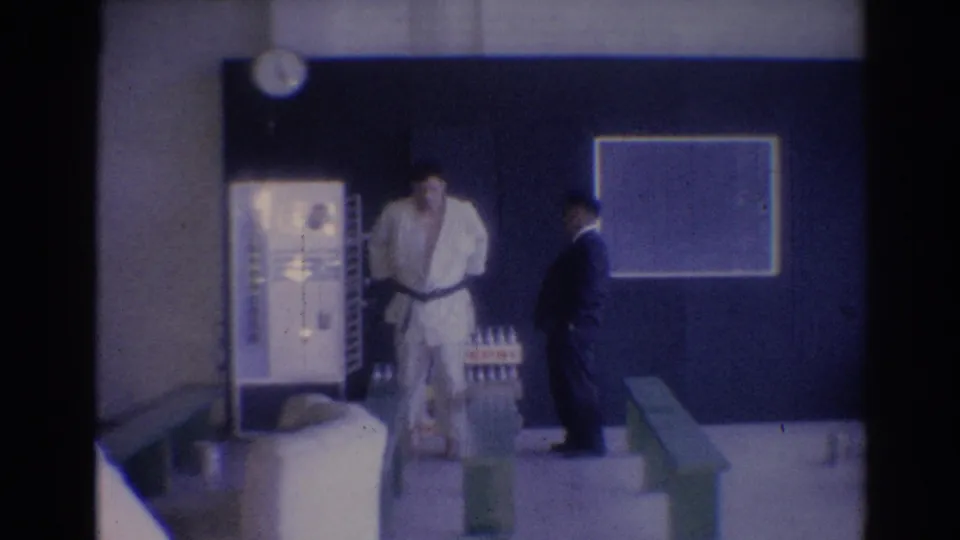 1967 Los Angeles gym frame with a judo-uniformed man near a vending machine, chalkboard, and suited man.
