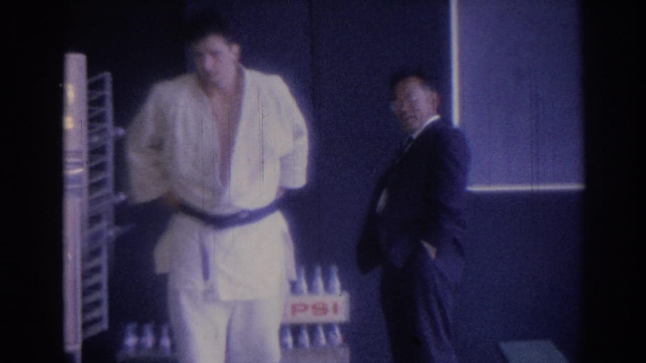 Martial artist in a white gi adjusts his belt while a suited man stands nearby.