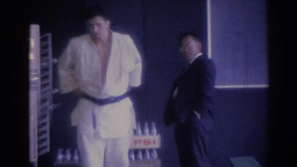 1967 Los Angeles home-movie frame showing a man in a white gi and black belt with a suited observer in a dim practice room.