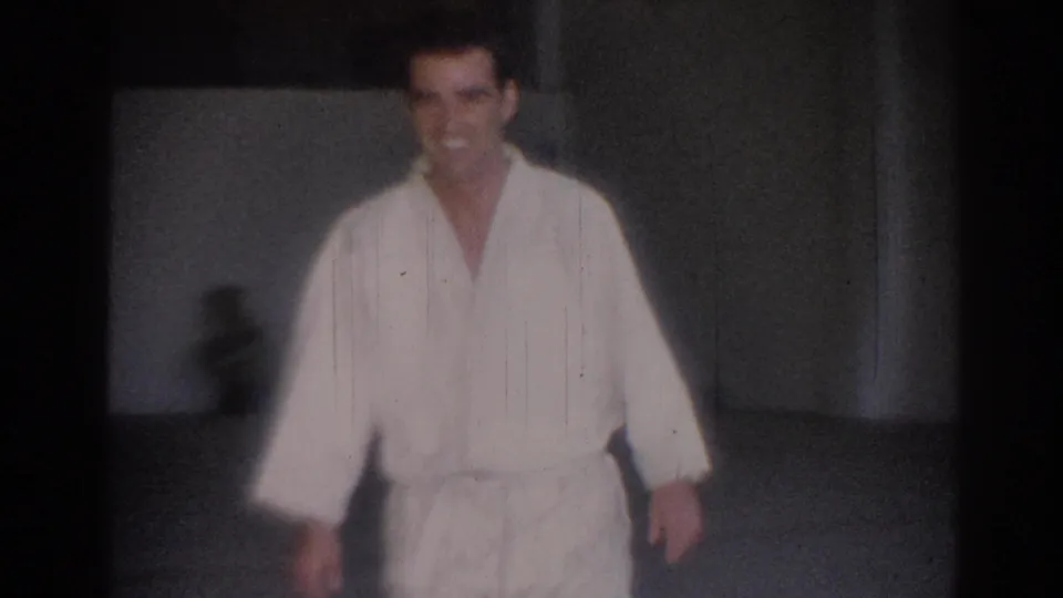 1967 Los Angeles home-movie frame showing a man in a white gi and black belt facing the camera in a sparse practice room.