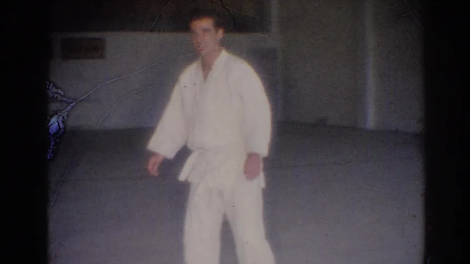 1967 Los Angeles frame showing a martial artist in a white gi and black belt walking across a dim practice-room floor.