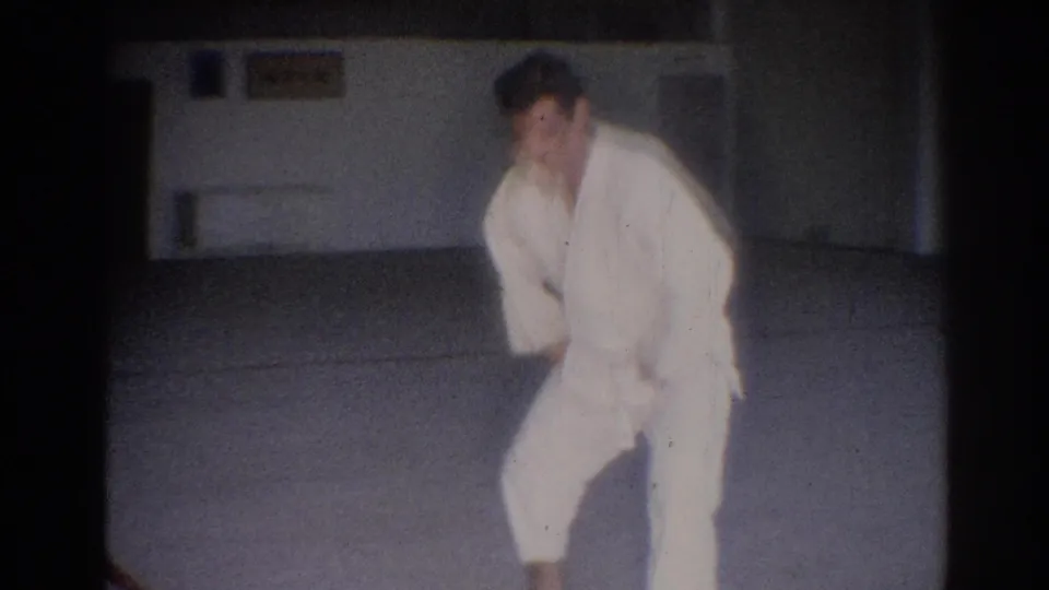 In 1967 Los Angeles home-movie footage, a man in a white judo gi bends into a stance on a pale mat inside a simple gym.