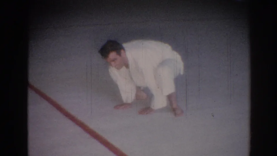 The judo practitioner crouches low on the mat near a red boundary line, with the soft blur and grain of 8mm film.