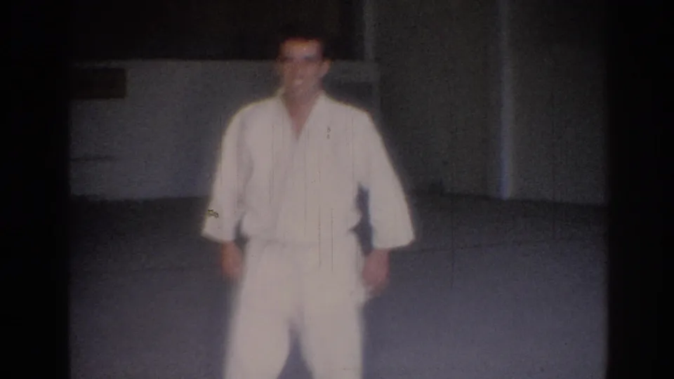 A man in a white judo uniform stands facing the camera in a simple Los Angeles gym setting, captured with soft 8mm focus.