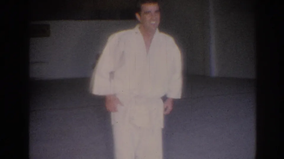 The performer stands in a white gi on the gym mat, framed by dark film edges and the faded color of 1960s 8mm footage.