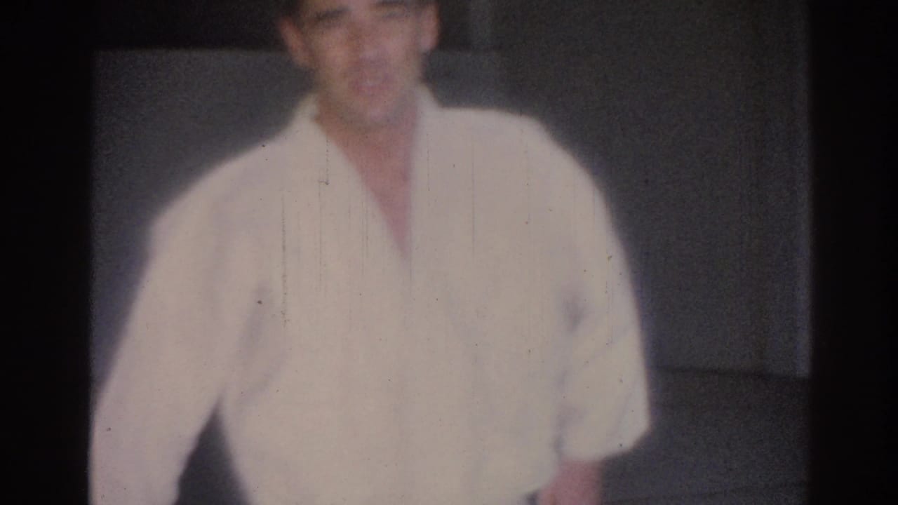 1967 Los Angeles home-movie frame showing a young man in a white judo gi facing the camera on an indoor mat.